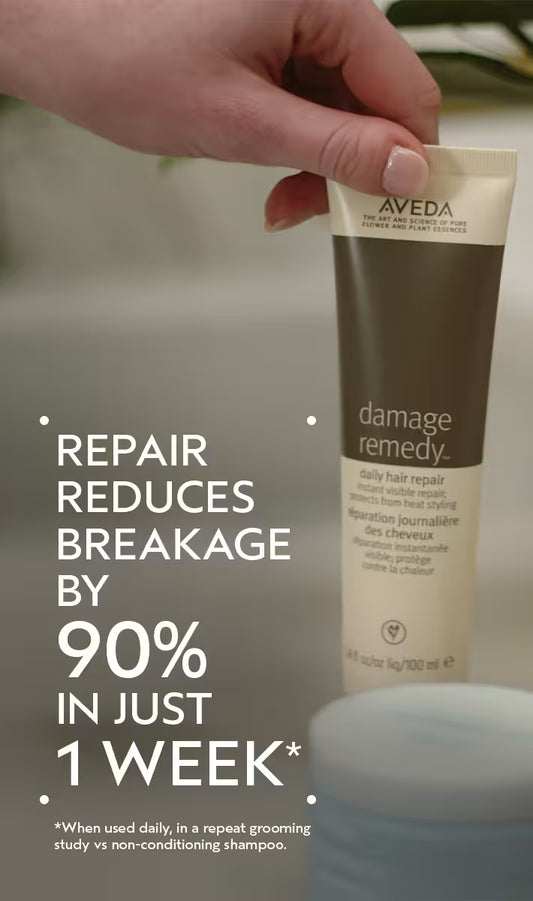 A hand holds an Aveda Damage Remedy hair repair tube against a softly blurred bathroom-like background, with large promotional text on the left reading 'REPAIR REDUCES BREAKAGE BY 90% IN JUST 1 WEEK'.