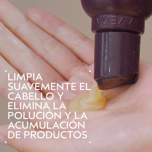 A close-up of a hand receiving a small swirl of yellow hair product from a dark brown pump bottle. White Spanish text overlays the left side, and the brand name Aveda is partially visible on the bottle.
